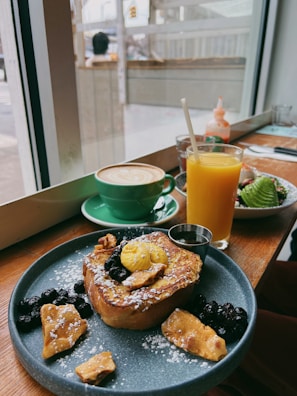 A cozy café setting featuring a wooden table with a delicious breakfast spread. The plate contains French toast topped with powdered sugar, berries, nuts, and a dollop of butter. Next to it, a bright orange juice and a green cup of cappuccino with latte art are placed. Through the window, a blurred view of the street is visible.