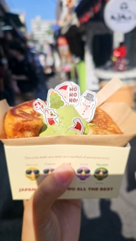 A hand is holding a cardboard box containing a dessert garnished with green matcha ice cream and several colorful Christmas-themed decorations. The background is a busy street with blurred shops and people, indicating a festive atmosphere.