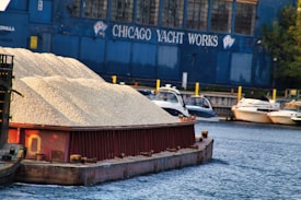 A barge loaded with gravel is floating on a river. Several yachts are docked at a marina behind the barge. The building in the background is painted blue with the words 'Chicago Yacht Works' visible. The scene captures a typical industrial and recreational waterfront setting.