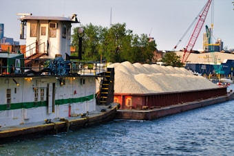 A barge loaded with a large pile of gravel is docked in a harbor. The vessel is white with a green stripe, and it's moored along a waterway with cables and equipment on its deck. In the background, there are trees, industrial buildings, and cranes, indicating a busy port area.