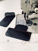 A sewing machine with a piece of dark fabric being stitched with red thread. Additional fabric pieces are placed nearby on a white surface, with visible red stitching and measuring lines in the background.