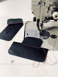 A sewing machine with a piece of dark fabric being stitched with red thread. Additional fabric pieces are placed nearby on a white surface, with visible red stitching and measuring lines in the background.