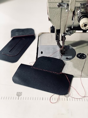 A sewing machine with a piece of dark fabric being stitched with red thread. Additional fabric pieces are placed nearby on a white surface, with visible red stitching and measuring lines in the background.