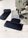 A sewing machine with a piece of dark fabric being stitched with red thread. Additional fabric pieces are placed nearby on a white surface, with visible red stitching and measuring lines in the background.