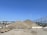 A construction site with a large mound of dirt, surrounded by concrete barriers and orange traffic cones. There is a clear blue sky overhead, and in the background, there are metal fences and distant hills.