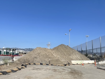 A construction site with a large mound of dirt, surrounded by concrete barriers and orange traffic cones. There is a clear blue sky overhead, and in the background, there are metal fences and distant hills.
