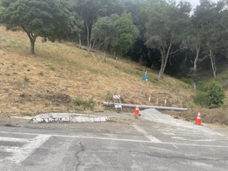 A sloped area of dry grass and several trees in the background, with a road at the forefront. Two construction cones and a barricade are present near a pipe and a small pile of gravel. A sign is also visible, mounted on a metal post.