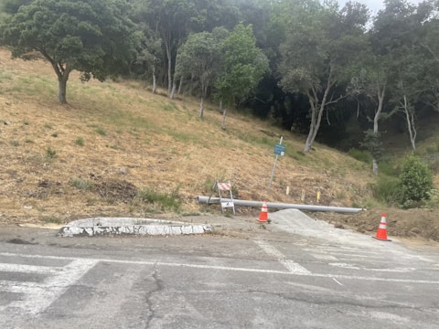 A sloped area of dry grass and several trees in the background, with a road at the forefront. Two construction cones and a barricade are present near a pipe and a small pile of gravel. A sign is also visible, mounted on a metal post.