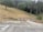 A sloped area of dry grass and several trees in the background, with a road at the forefront. Two construction cones and a barricade are present near a pipe and a small pile of gravel. A sign is also visible, mounted on a metal post.