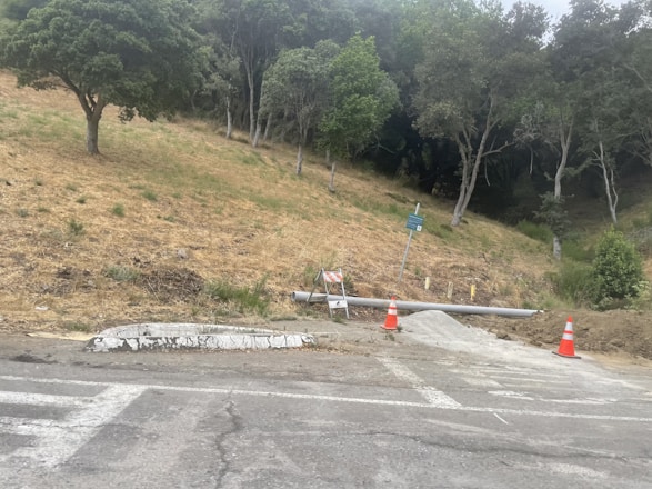 A sloped area of dry grass and several trees in the background, with a road at the forefront. Two construction cones and a barricade are present near a pipe and a small pile of gravel. A sign is also visible, mounted on a metal post.