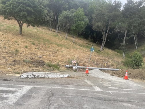A sloped area of dry grass and several trees in the background, with a road at the forefront. Two construction cones and a barricade are present near a pipe and a small pile of gravel. A sign is also visible, mounted on a metal post.