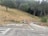 A sloped area of dry grass and several trees in the background, with a road at the forefront. Two construction cones and a barricade are present near a pipe and a small pile of gravel. A sign is also visible, mounted on a metal post.