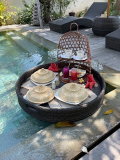 A luxurious floating breakfast setup is placed in a pool. The round wicker tray holds plates covered with woven lids, cups of coffee, and vibrant juice glasses. There are red napkins folded elegantly, accompanied by small flowers for decoration. The background features wooden decking and two black wicker lounge chairs, surrounded by greenery.