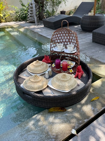 A luxurious floating breakfast setup is placed in a pool. The round wicker tray holds plates covered with woven lids, cups of coffee, and vibrant juice glasses. There are red napkins folded elegantly, accompanied by small flowers for decoration. The background features wooden decking and two black wicker lounge chairs, surrounded by greenery.