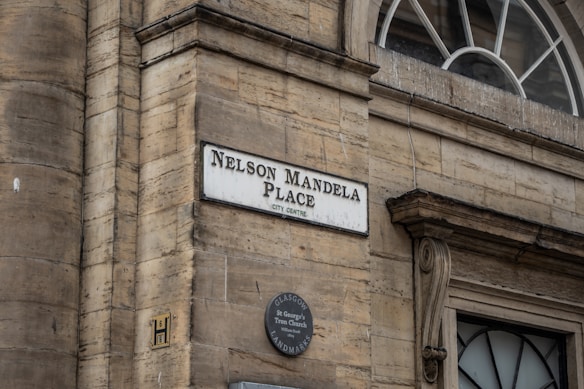 A street sign mounted on a stone wall indicates 'Nelson Mandela Place, City Centre'. Below the street sign, a round plaque reads 'Glasgow St George's Tron Church'. An ornate architectural element frames a window with a semi-circular top.