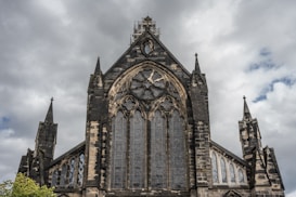 The image features a gothic-style church facade with intricate stonework and large stained glass windows. The architecture includes pointed arches and spires reaching toward a cloudy sky, adding a dramatic and historic feel.