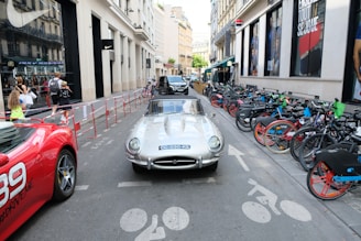 A silver vintage convertible car is parked in a busy city street surrounded by a red sports car on the left and a line of bicycles on the right. Pedestrians, including adults and children, walk past stores on the left side, and various advertisements are visible on the buildings.