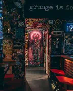 A hallway filled with vibrant graffiti and street art, leading to a striking mural depicting a person with a guitar. The walls are covered in various layers of colorful spray paint and stickers. Neon lighting adds a dramatic effect to the scene.