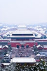 Snow-covered traditional Chinese architecture extends into the distance, creating a symmetrical and serene view of an ancient palace complex. The roofs and trees are lightly dusted with snow, adding a wintery atmosphere. Visitors can be seen walking through the complex's entrance, suggesting an active and engaging environment.