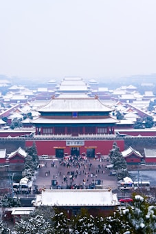 Snow-covered traditional Chinese architecture extends into the distance, creating a symmetrical and serene view of an ancient palace complex. The roofs and trees are lightly dusted with snow, adding a wintery atmosphere. Visitors can be seen walking through the complex's entrance, suggesting an active and engaging environment.