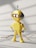A small, yellow robot toy stands on a light-colored surface with its round head featuring two glowing eyes and a small antenna. The robot has rounded limbs and an oval panel on its chest. Shadows and a stray stick or twig are visible on the ground.