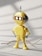 A small, yellow robot toy stands on a light-colored surface with its round head featuring two glowing eyes and a small antenna. The robot has rounded limbs and an oval panel on its chest. Shadows and a stray stick or twig are visible on the ground.