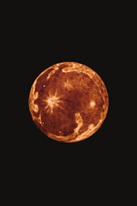 A spherical celestial body with a textured, mottled surface in shades of brown and orange against a dark background. Bright spots and lines radiate in some areas, resembling craters or impact points.