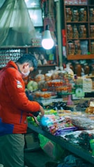 A person in a red jacket and face mask stands at a market stall, examining packaged goods on a counter. The stall is lit by a single hanging light bulb, and various jars and products are displayed in the background.