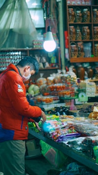 A person in a red jacket and face mask stands at a market stall, examining packaged goods on a counter. The stall is lit by a single hanging light bulb, and various jars and products are displayed in the background.
