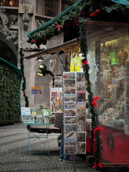 A festive market stall decorated with garlands and red bows, displaying various postcards and souvenirs. The stall is adorned with greenery and brightly colored ornaments, creating a warm and inviting holiday atmosphere. The cobblestone pavement and ornate old building in the background suggest a historical setting.