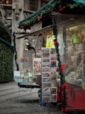 A festive market stall decorated with garlands and red bows, displaying various postcards and souvenirs. The stall is adorned with greenery and brightly colored ornaments, creating a warm and inviting holiday atmosphere. The cobblestone pavement and ornate old building in the background suggest a historical setting.