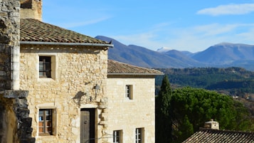 A rustic stone house with a tiled roof, featuring wooden windows and a door, set against a backdrop of mountains and lush green trees. The foreground shows part of another stone wall with a lantern.