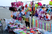 A vibrant market stall on a beach displays an array of colorful toys and accessories. Items include stuffed toys, hats, caps, plastic toys, and decorative ribbons. The background shows a sandy beach with people and parasols.