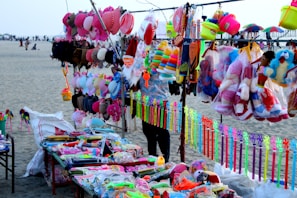 A vibrant market stall on a beach displays an array of colorful toys and accessories. Items include stuffed toys, hats, caps, plastic toys, and decorative ribbons. The background shows a sandy beach with people and parasols.