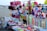 A vibrant market stall on a beach displays an array of colorful toys and accessories. Items include stuffed toys, hats, caps, plastic toys, and decorative ribbons. The background shows a sandy beach with people and parasols.