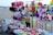 A vibrant market stall on a beach displays an array of colorful toys and accessories. Items include stuffed toys, hats, caps, plastic toys, and decorative ribbons. The background shows a sandy beach with people and parasols.