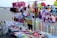 A vibrant market stall on a beach displays an array of colorful toys and accessories. Items include stuffed toys, hats, caps, plastic toys, and decorative ribbons. The background shows a sandy beach with people and parasols.