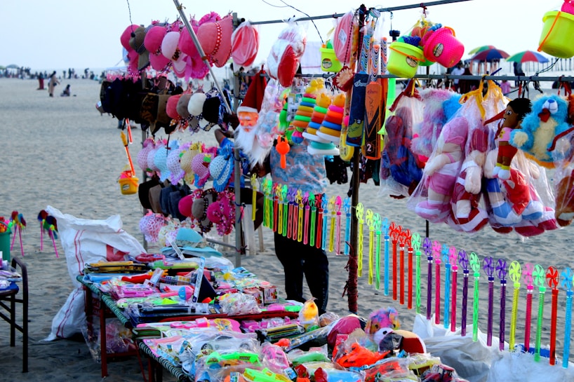 A vibrant market stall on a beach displays an array of colorful toys and accessories. Items include stuffed toys, hats, caps, plastic toys, and decorative ribbons. The background shows a sandy beach with people and parasols.