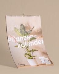 A spiral-bound calendar featuring a large plant in a white pot at the center. The background is a neutral beige color, and the words 'immer-währender Pflanzenkalender' are prominently displayed in white text. There are two human arms extending into the frame, suggesting a sense of interaction with the plant. At the bottom of the calendar, the brand name 'feey' is visible, and a couple of brochure-like pages are positioned on the right side.