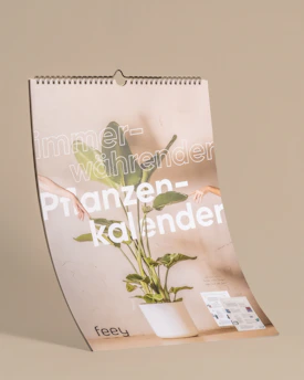 A spiral-bound calendar featuring a large plant in a white pot at the center. The background is a neutral beige color, and the words 'immer-währender Pflanzenkalender' are prominently displayed in white text. There are two human arms extending into the frame, suggesting a sense of interaction with the plant. At the bottom of the calendar, the brand name 'feey' is visible, and a couple of brochure-like pages are positioned on the right side.