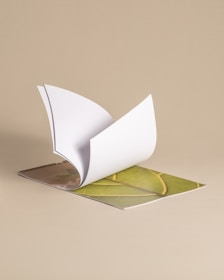 A spiral-bound book with a thick cover partially open and flipped to reveal a page with a vivid green leaf design. The background is a plain, soft beige.
