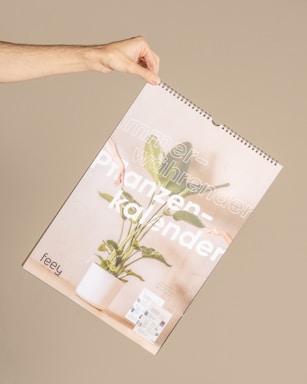 A hand holding up a wall calendar featuring a potted plant with green leaves. The calendar has large text in a light font that partially reads 'immerwährender Pflanzenkalender.' The background is a muted beige color, complementing the soft, neutral tones of the calendar.