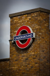A prominent, circular sign with a red outer ring and blue rectangular bar is mounted on a brick wall. The word 'UNDERGROUND' is displayed in white letters across the blue bar. The brick wall is made of yellowish-brown bricks, and the sky above is overcast, adding a moody atmosphere.