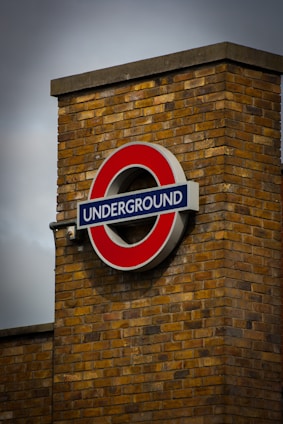 A prominent, circular sign with a red outer ring and blue rectangular bar is mounted on a brick wall. The word 'UNDERGROUND' is displayed in white letters across the blue bar. The brick wall is made of yellowish-brown bricks, and the sky above is overcast, adding a moody atmosphere.