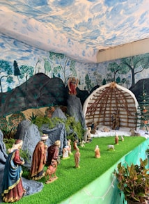 A nativity scene is displayed with various figurines representing biblical characters on a green, grassy platform. The backdrop is painted with a sky and forest landscape, creating a serene setting. An angel figure is placed on a rock formation, overseeing the scene. A stable structure is featured, housing more figurines, including shepherds and animals.