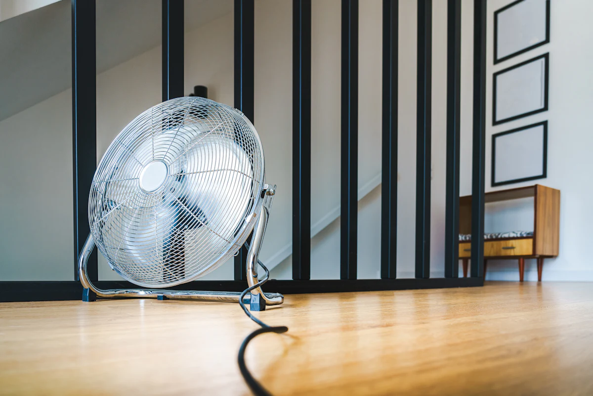 Ventilador turbo metálico apoyado en el piso de madera de un living, junto a una baranda