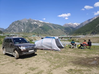 A group of people are camping in a scenic mountainous area. There is a grey and blue tent set up on a grassy area near a river. Three individuals are sitting on camping chairs around a table, engaging in an outdoor activity. A black SUV with a visible license plate is parked nearby on the grass. The background features snow-capped mountains, green hills, trees, scattered houses, and a clear blue sky with a few clouds.