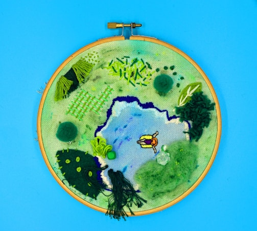An embroidery hoop art piece displays a landscape scene with a mix of thread textures and colors. The scene includes a central pond with blue and purple hues, surrounded by green foliage and grass. A small figure dressed in orange lies near the water. The background is a vivid blue, likely representing the sky.