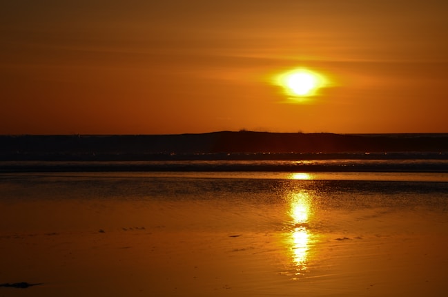 A serene beach scene at sunset with the sun low in the sky casting a warm golden glow. The light reflects off the calm water in the foreground, creating a peaceful and tranquil atmosphere.