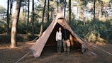 Two-person camping tent pitched in a lush forest clearing at sunrise.
